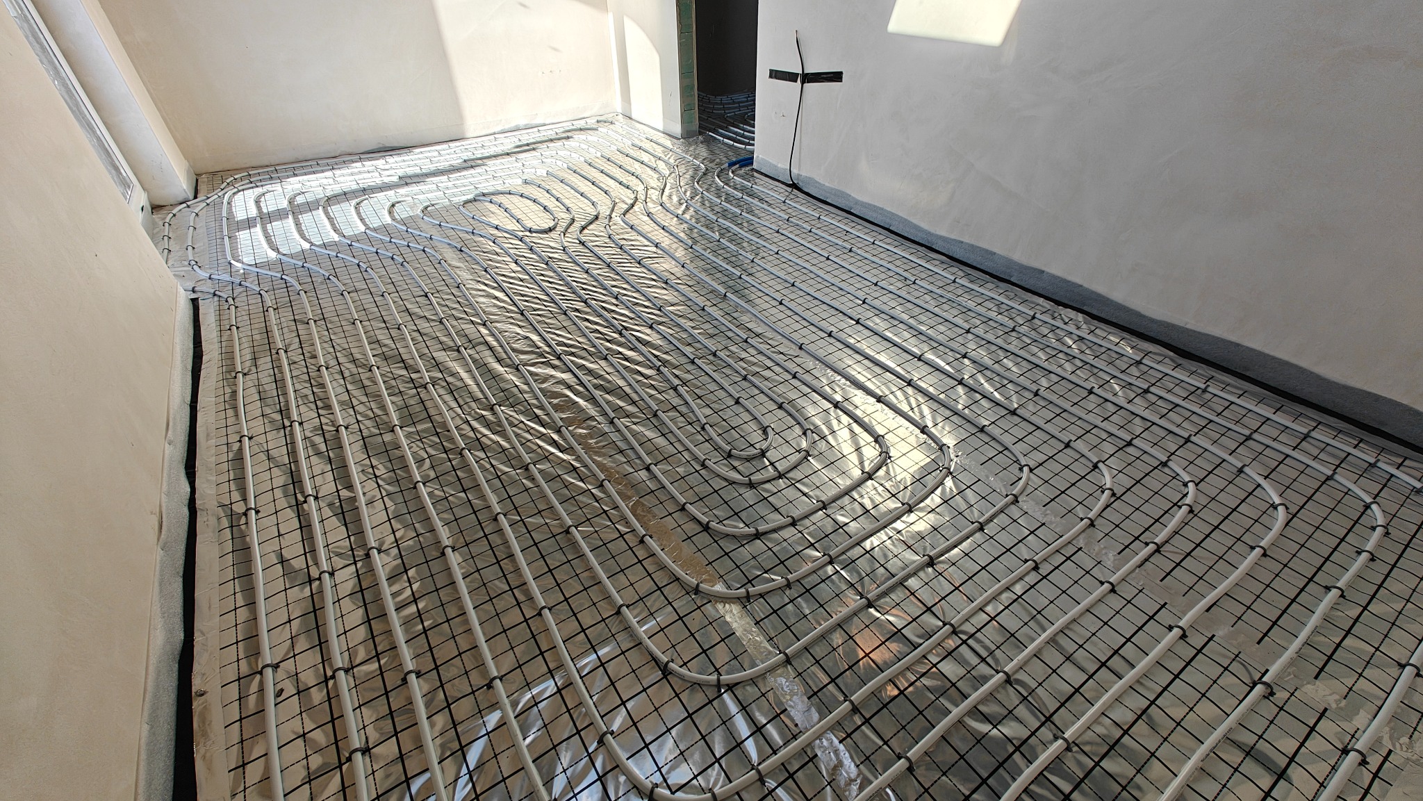 Białe rury ogrzewania podłogowego ułożone na folii izolacyjnej w jasnym pomieszczeniu. Widoczne mocowania rur do siatki. Ściany w surowym stanie.