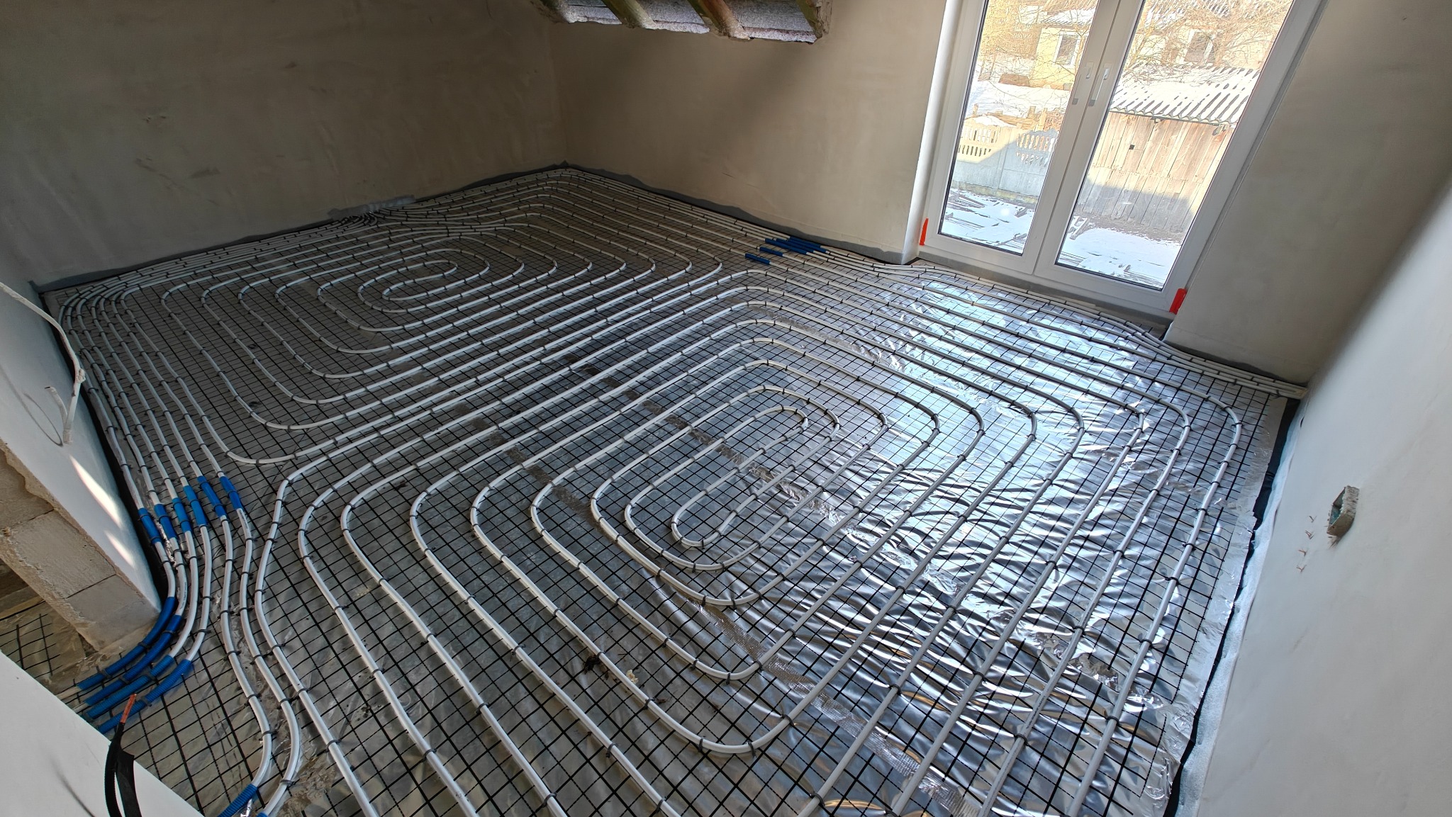 Białe rury ogrzewania podłogowego ułożone spiralnie na folii izolacyjnej w surowym pomieszczeniu z oknem. Niebieskie złączki na końcach rur.