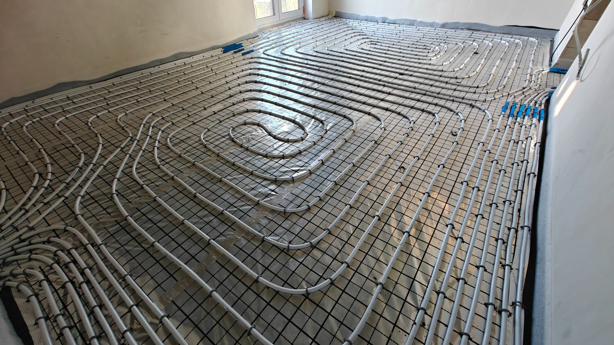 Białe rury ogrzewania podłogowego ułożone spiralnie na czarnej siatce, widoczne niebieskie złączki, przygotowane do zalania wylewką w jasnym pomieszczeniu.