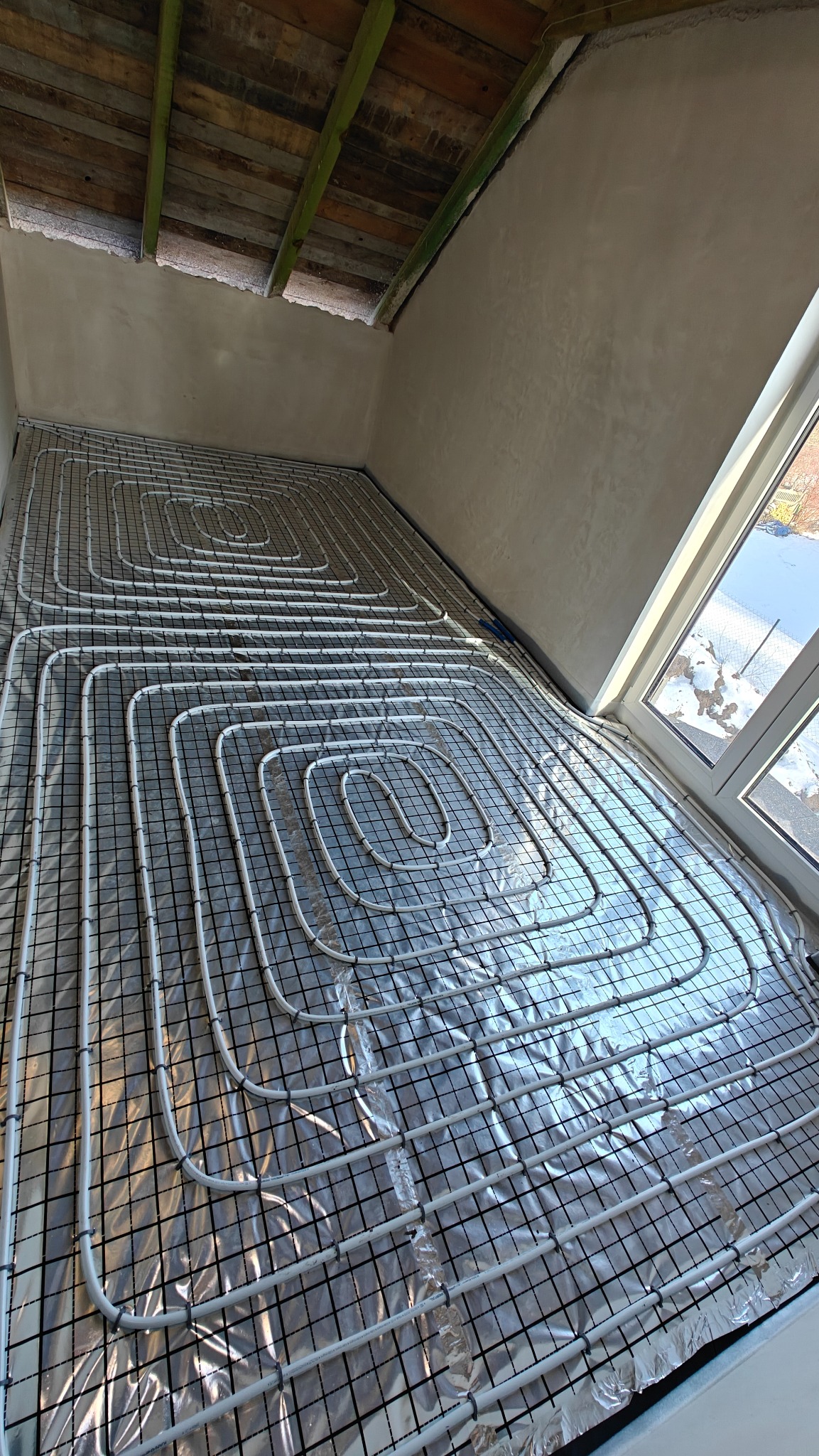 Białe rury ogrzewania podłogowego ułożone na folii w Lucynowie, widok z góry. Instalacja przed wylaniem posadzki, okno w tle.