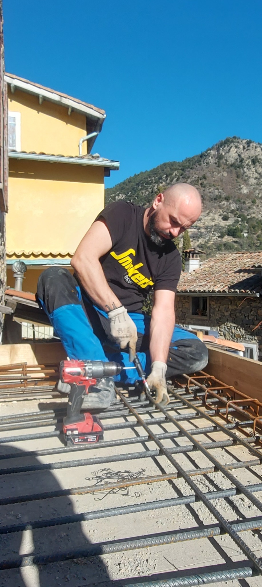 Pracownik w pozycji kucznej wiąże zbrojenie na tle żółtego budynku i gór. W pobliżu leży wiertarko-wkrętarka. Zastosowano jasne, naturalne oświetlenie.