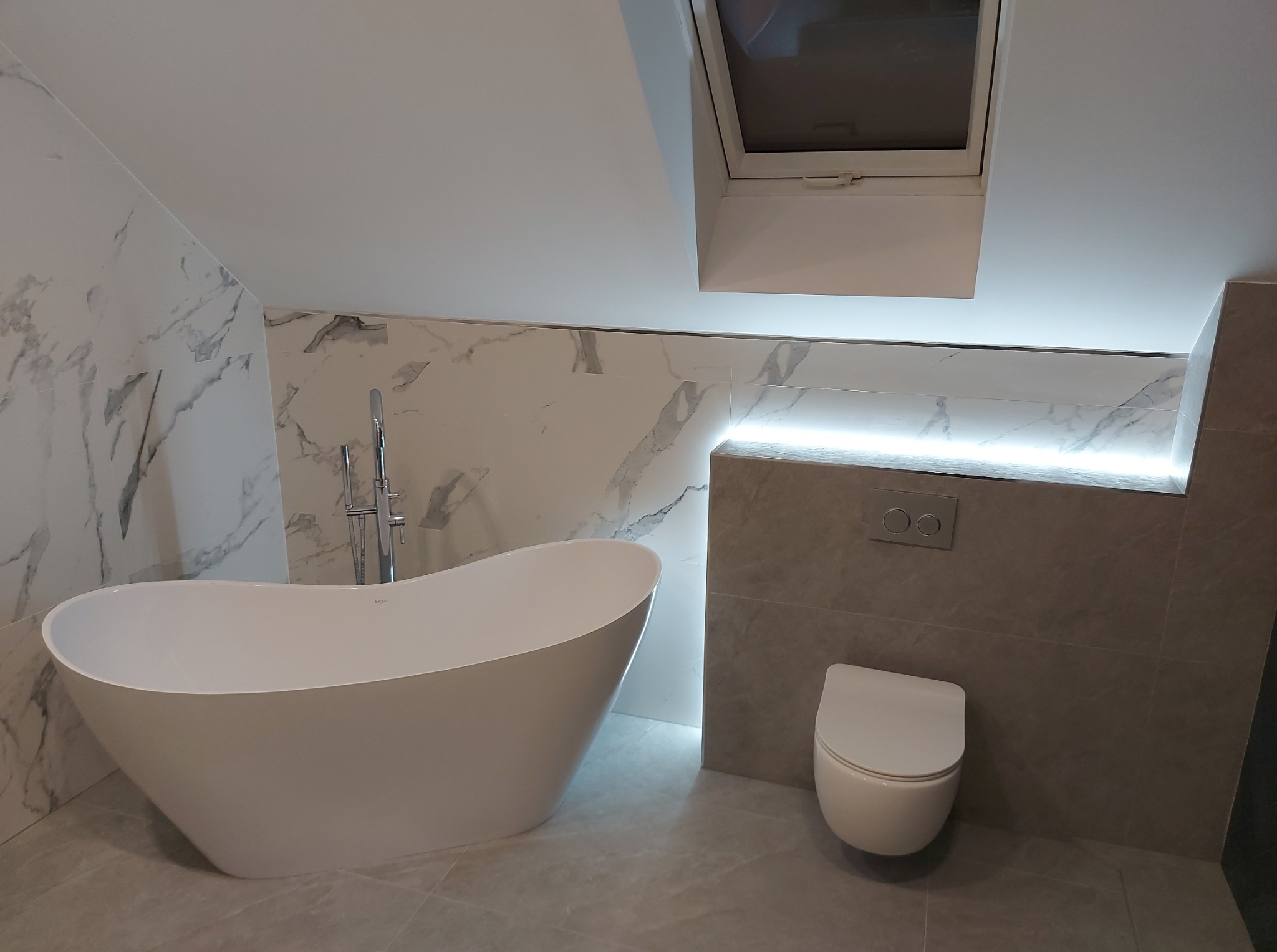 Nowoczesna łazienka na poddaszu z wolnostojącą wanną, marmurowymi płytkami i podświetleniem LED. Uwagę zwraca okno dachowe wkomponowane w skos.