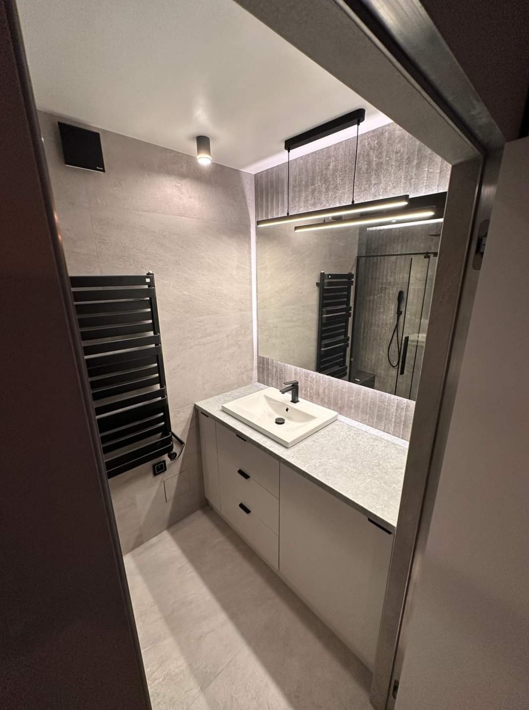 Nowoczesna łazienka z umywalką nablatową, grzejnikiem drabinkowym i podświetlanym lustrem. Minimalistyczny design, szare płytki i białe meble.