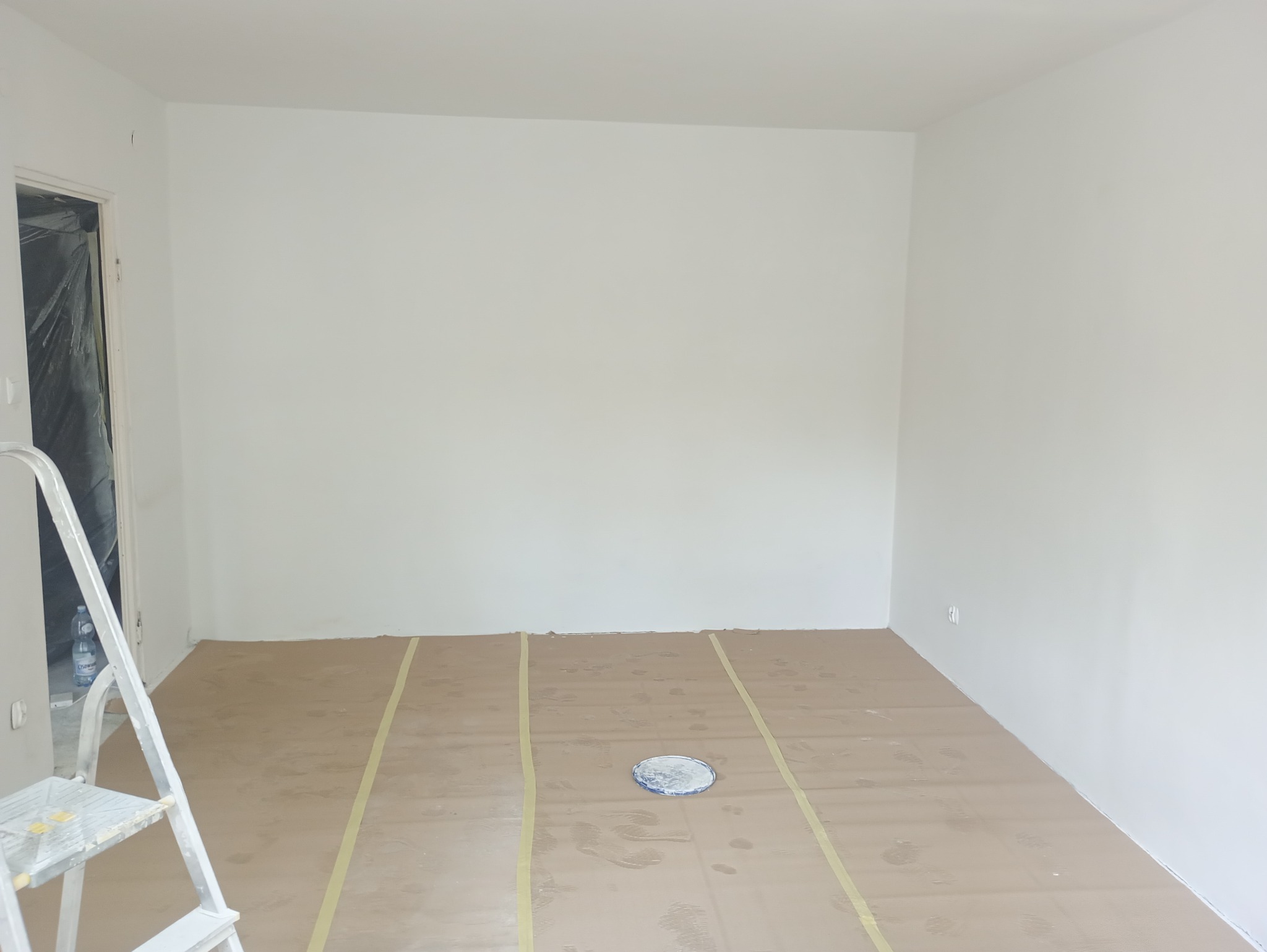 Białe ściany po malowaniu w pokoju, podłoga zabezpieczona kartonem i taśmą malarską. W rogu drabina malarska. Widok na świeżo pomalowane wnętrze.