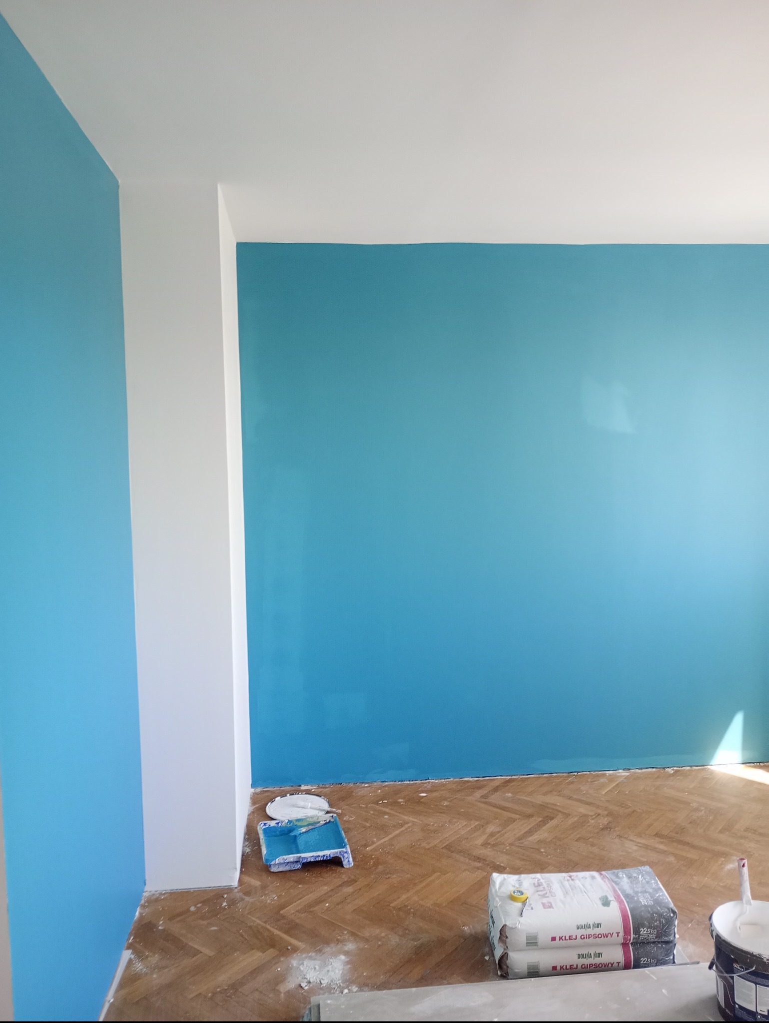 Turkusowa ściana po malowaniu z widocznymi narzędziami: wałek, kuweta, klej gipsowy na parkiecie. Świeżo pomalowane ściany w pokoju.