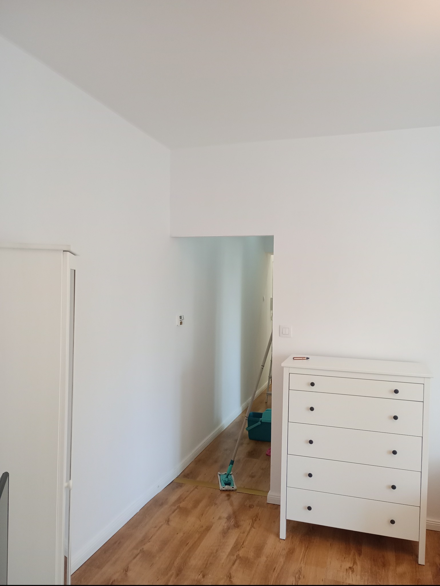 Białe, świeżo pomalowane ściany w pokoju z drewnianą podłogą. Widoczna drabina malarska w przejściu i biała komoda z ciemnymi uchwytami.
