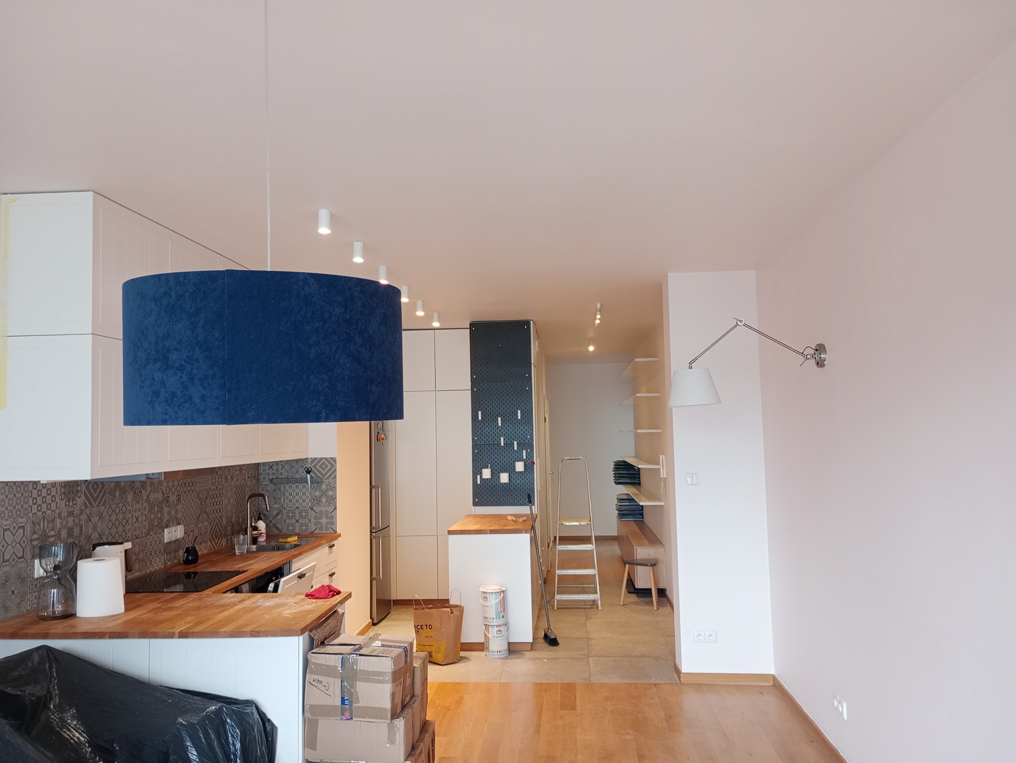 Wnętrze mieszkania w trakcie odświeżania: pomalowane ściany, zabezpieczone meble, drabina w tle. Widoczne puszki z farbą i akcesoria malarskie.