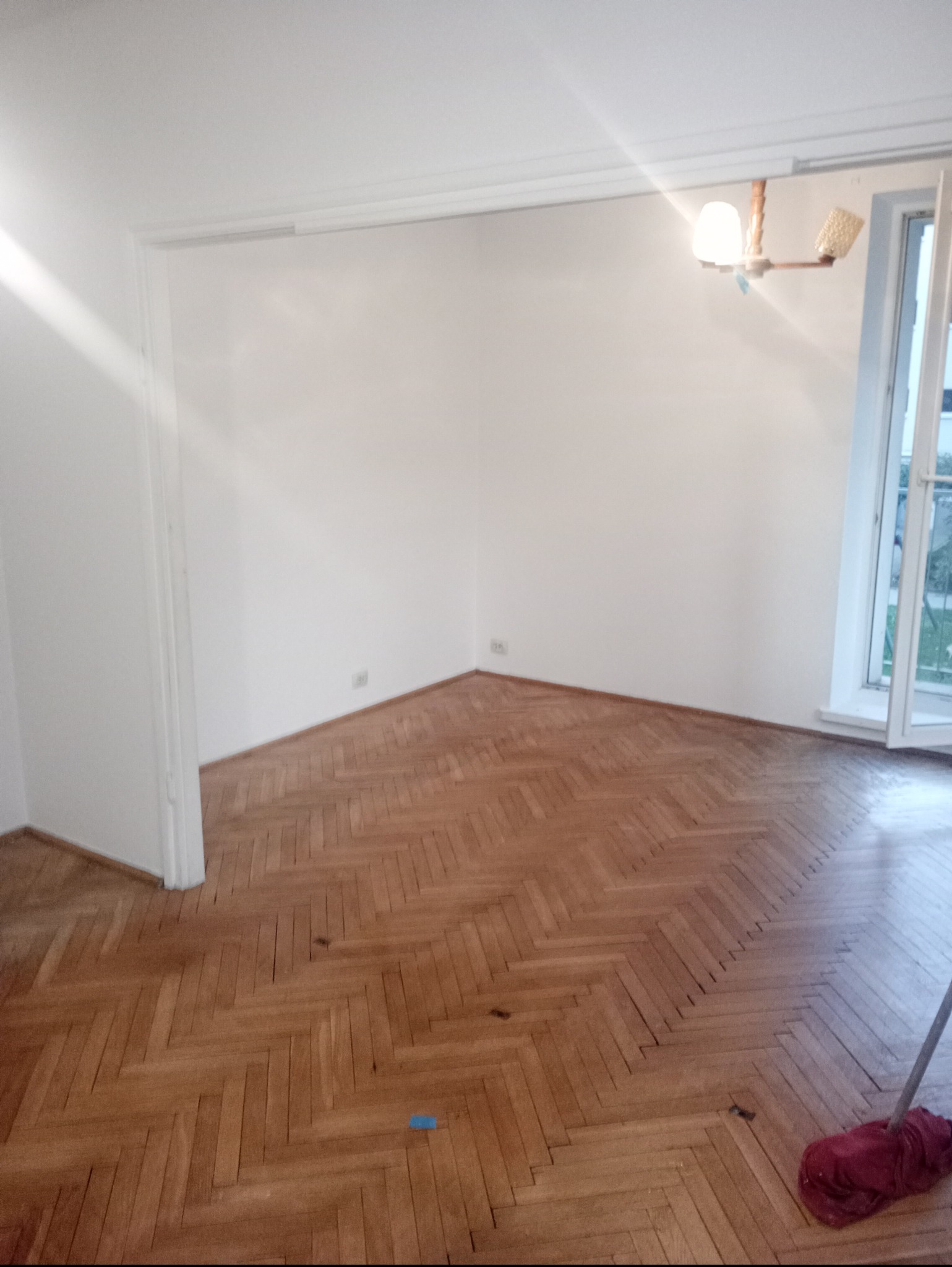 Jasne wnętrze po malowaniu: białe ściany, parkiet w jodełkę, otwarte okno z widokiem na zieleń. W prawym dolnym rogu widoczny mop.