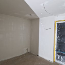 Tomasz Lisek - Świeżo pomalowane, jasne wnętrze z zabezpieczonymi folią oknami. Widoczne punkty oświetleniowe w suficie i wystające przewody. Ściany gładkie, gotowe do dalszych prac.