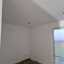 Tomasz Lisek - Świeżo pomalowane na biało wnętrze pokoju z oknem zabezpieczonym folią. Widoczne otwory na oświetlenie w suficie i listwy przypodłogowe.