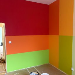 Tomasz Lisek - Wnętrze po malowaniu w pasy: czerwony, pomarańczowy, zielony. Widoczne puszki z farbą i taśmy malarskie na podłodze. Świeżo pomalowane ściany w żywych kolorach.