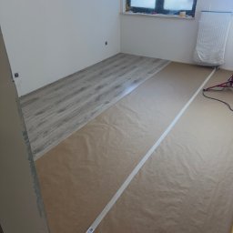 Tomasz Lisek - Układanie paneli podłogowych w pokoju, część podłogi zabezpieczona papierem malarskim. Widoczne narzędzia i okno z osłoniętym grzejnikiem.