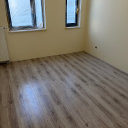 Tomasz Lisek - Pusty pokój po remoncie z nową podłogą z paneli i zabezpieczonymi oknami folią. Ściany pomalowane na jasny kolor. Widoczny grzejnik i gniazdko elektryczne.
