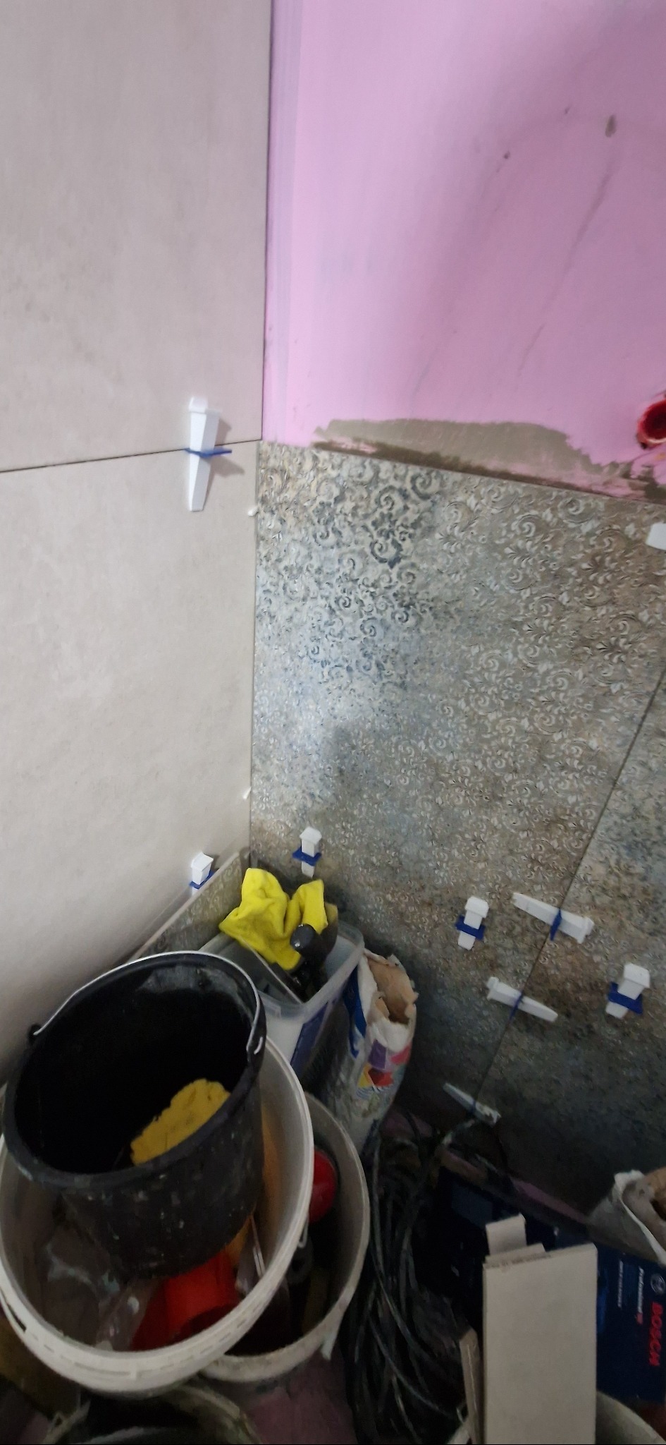 Układanie płytek w łazience z użyciem krzyżyków dystansowych. Widoczne narzędzia i materiały budowlane. Ściana pomalowana na różowo w górnym rogu.