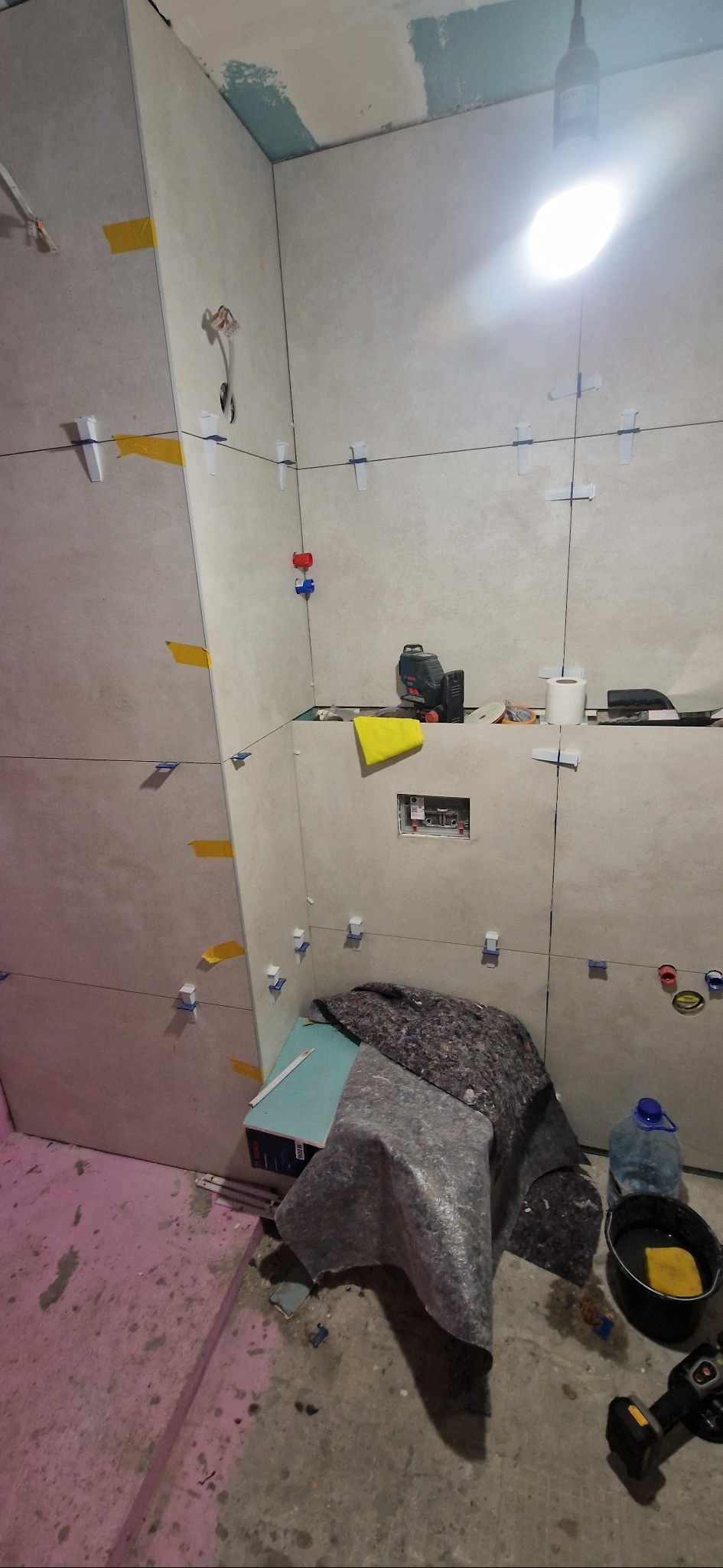 Wnętrze łazienki w trakcie prac glazurniczych: ułożone płytki z użyciem krzyżyków dystansowych, poziomica laserowa, narzędzia i materiały budowlane na podłodze.