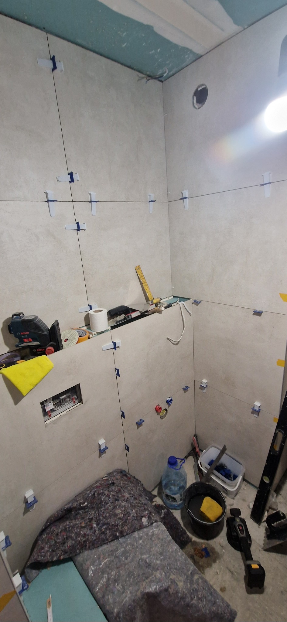 Układanie płytek w łazience, widoczne krzyżyki dystansowe, poziomica, laserowy niwelator i inne narzędzia glazurnicze. Ściany wyłożone jasnymi płytkami.