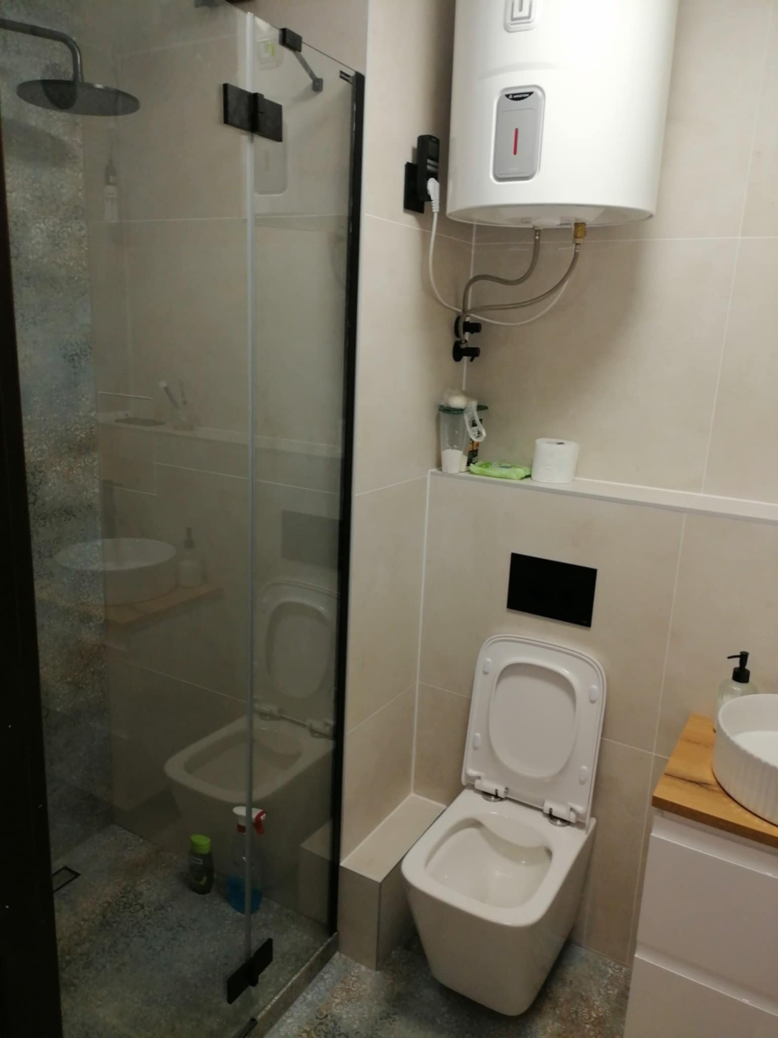 Nowoczesna łazienka z kabiną prysznicową typu walk-in, podwieszaną toaletą i bojlerem. Beżowe płytki, czarne akcenty. Umywalka nablatowa na drewnianym blacie.