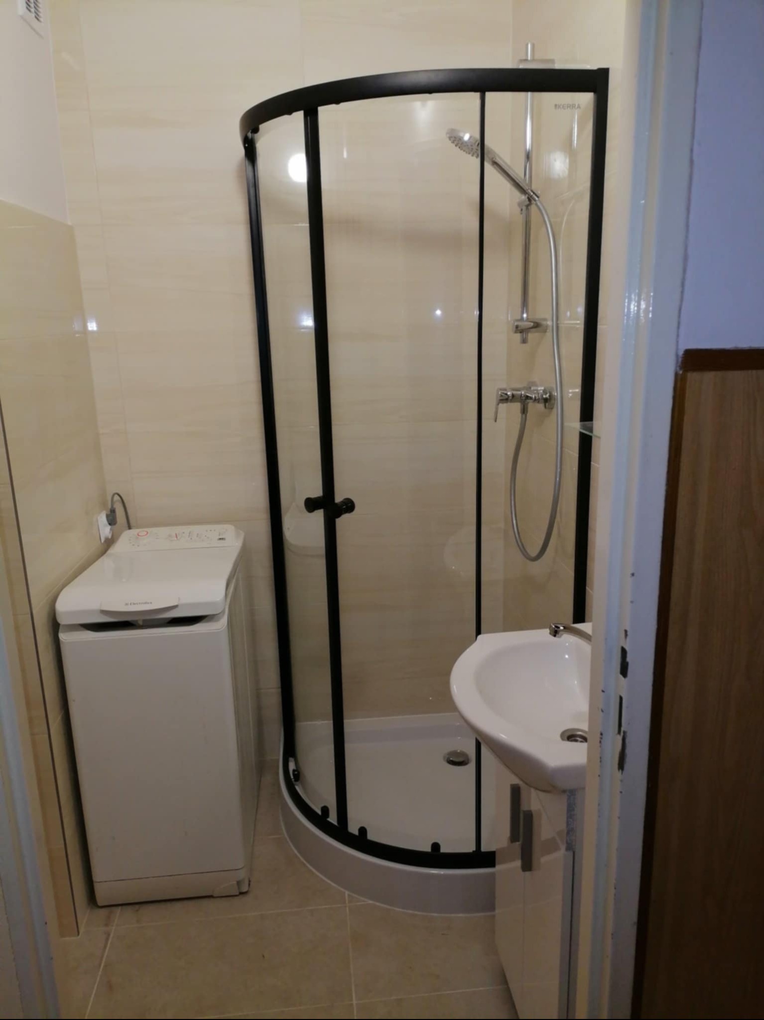 Jasna łazienka po remoncie: kabina prysznicowa z czarnymi ramami, umywalka z szafką i pralka. Beżowe płytki na ścianach i podłodze. Widok zza futryny.