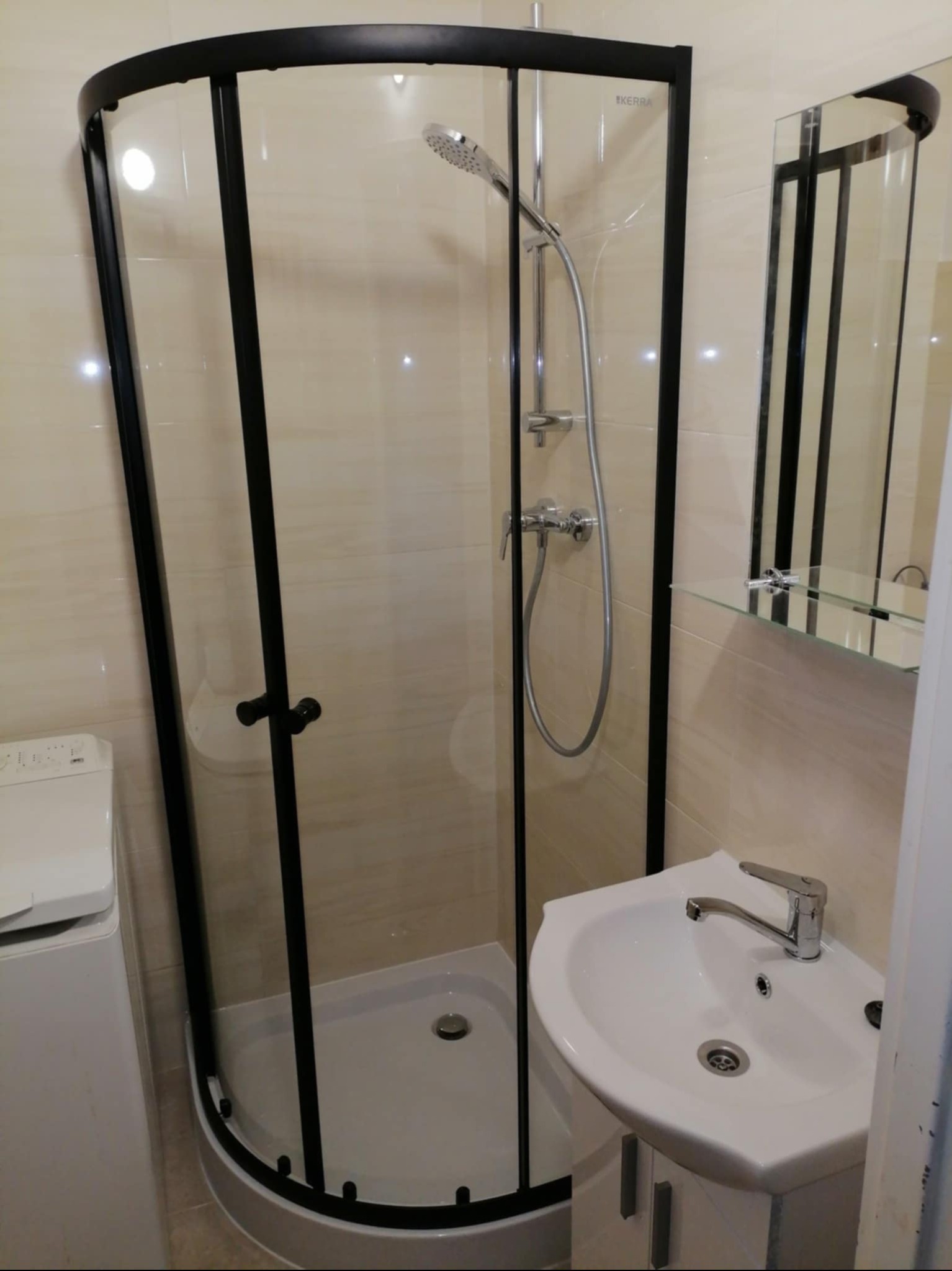 Nowoczesna kabina prysznicowa z czarnymi profilami, umywalka z szafką i pralka w jasnej łazience wykończonej beżowymi płytkami. Widoczne lustro z półką.