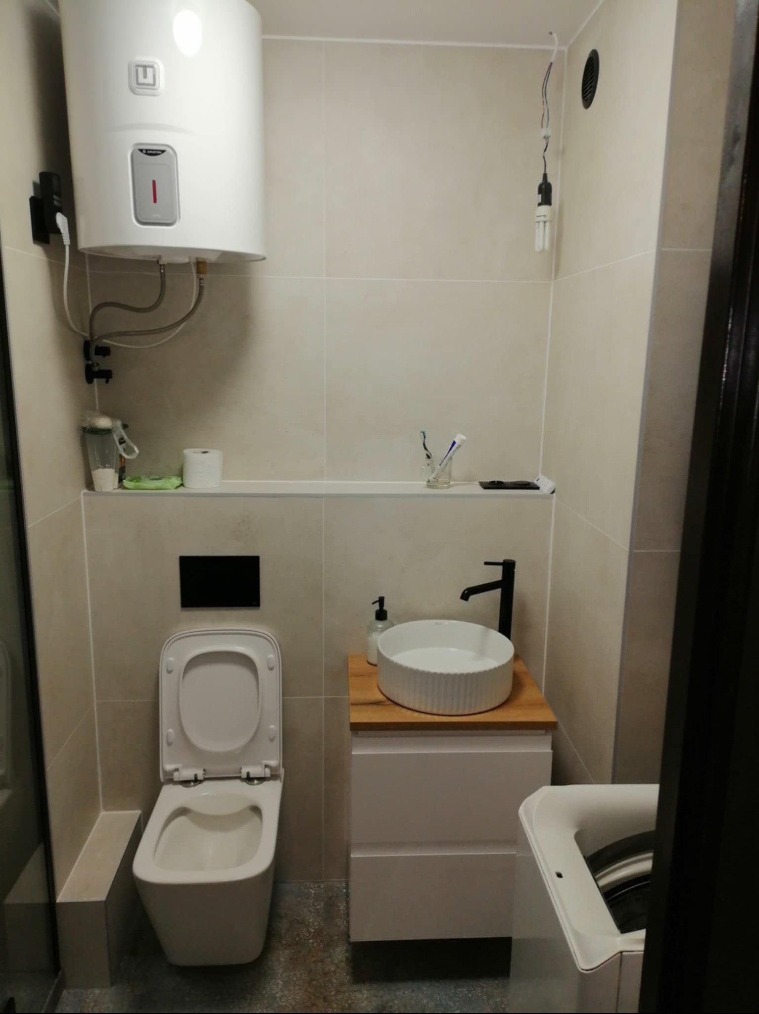 Nowoczesna łazienka po remoncie: beżowe płytki, podwieszana toaleta, biała umywalka na drewnianym blacie, bojler i pralka. Minimalistyczny design.