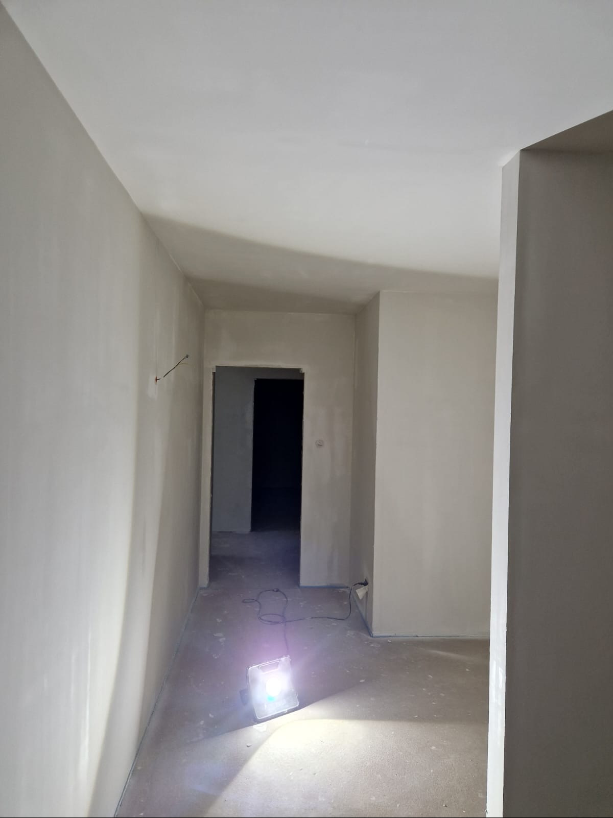 Wnętrze po gładziowaniu ścian, widoczny halogen budowlany na podłodze, przygotowane do malowania. Kabel elektryczny wystaje ze ściany.