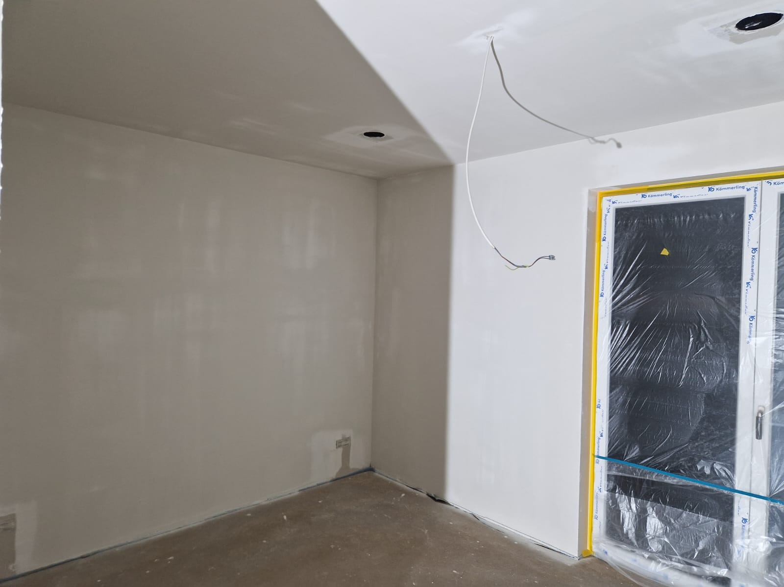 Świeżo pomalowane, jasne wnętrze z zabezpieczonymi folią oknami. Widoczne punkty oświetleniowe w suficie i wystające przewody. Ściany gładkie, gotowe do dalszych prac.