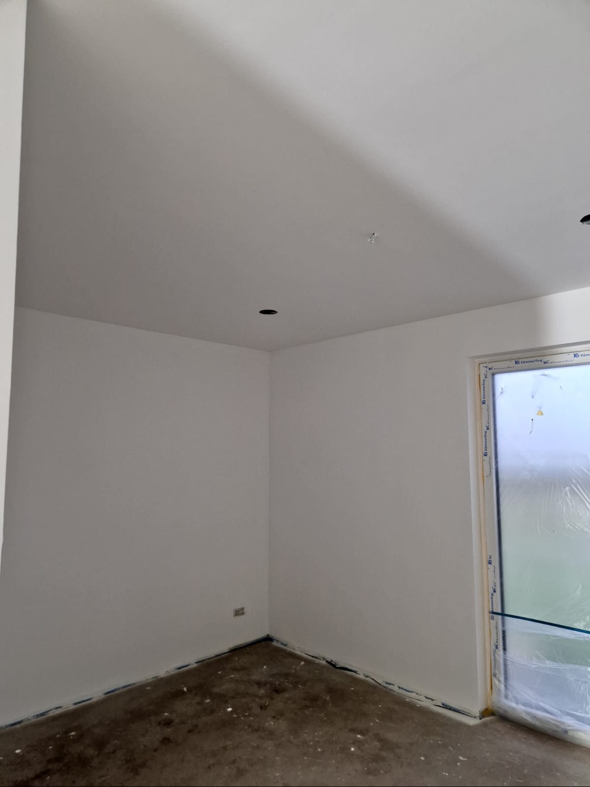 Świeżo pomalowane na biało wnętrze pokoju z oknem zabezpieczonym folią. Widoczne otwory na oświetlenie w suficie i listwy przypodłogowe.