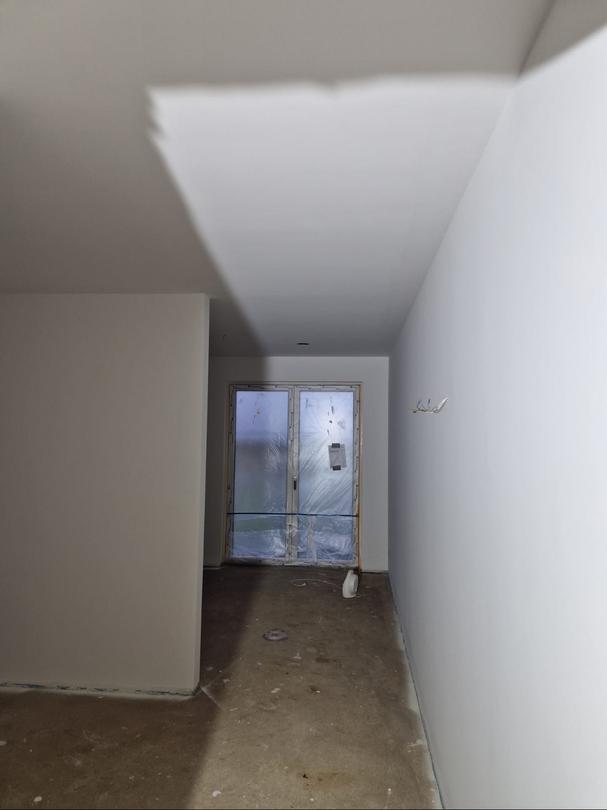 Wnętrze w trakcie malowania: świeżo pomalowane ściany na biało, okna zabezpieczone folią malarską, surowa podłoga. Widoczne ślady po pracach malarskich.