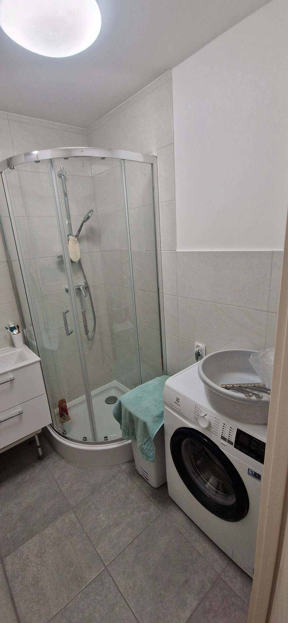 Jasna łazienka po remoncie: kabina prysznicowa, umywalka z szafką i pralka Electrolux z miską na wierzchu. Szare płytki podłogowe i ścienne.