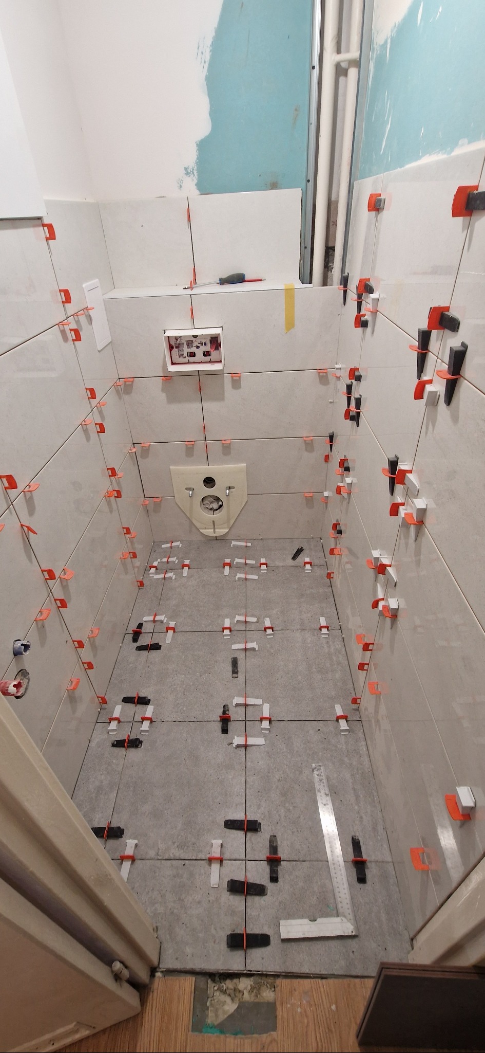 Łazienka w trakcie układania płytek podłogowych i ściennych z użyciem klipsów poziomujących. Widoczny fragment ściany z instalacją sanitarną i narzędzia glazurnicze.