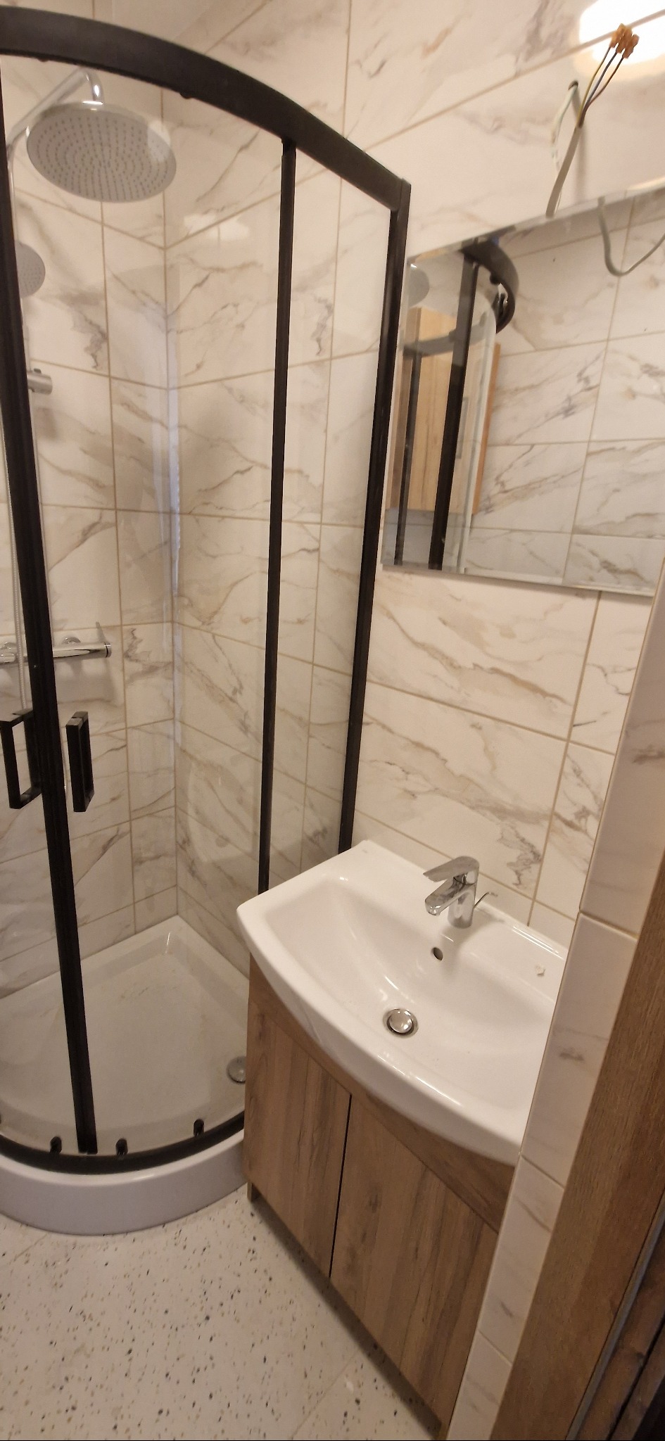 Nowoczesna kabina prysznicowa z czarną ramą i umywalka z szafką w wykończeniu drewnopodobnym, marmurowe płytki na ścianach, lustro w tle.