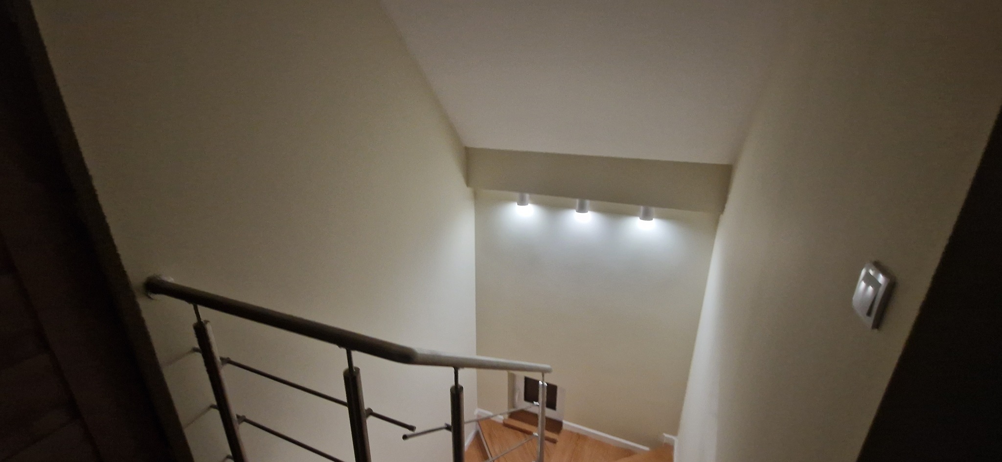 Jasne wnętrze klatki schodowej z drewnianymi schodami i metalową balustradą. Ściany w odcieniach beżu, oświetlenie punktowe na suficie. Widok z góry.