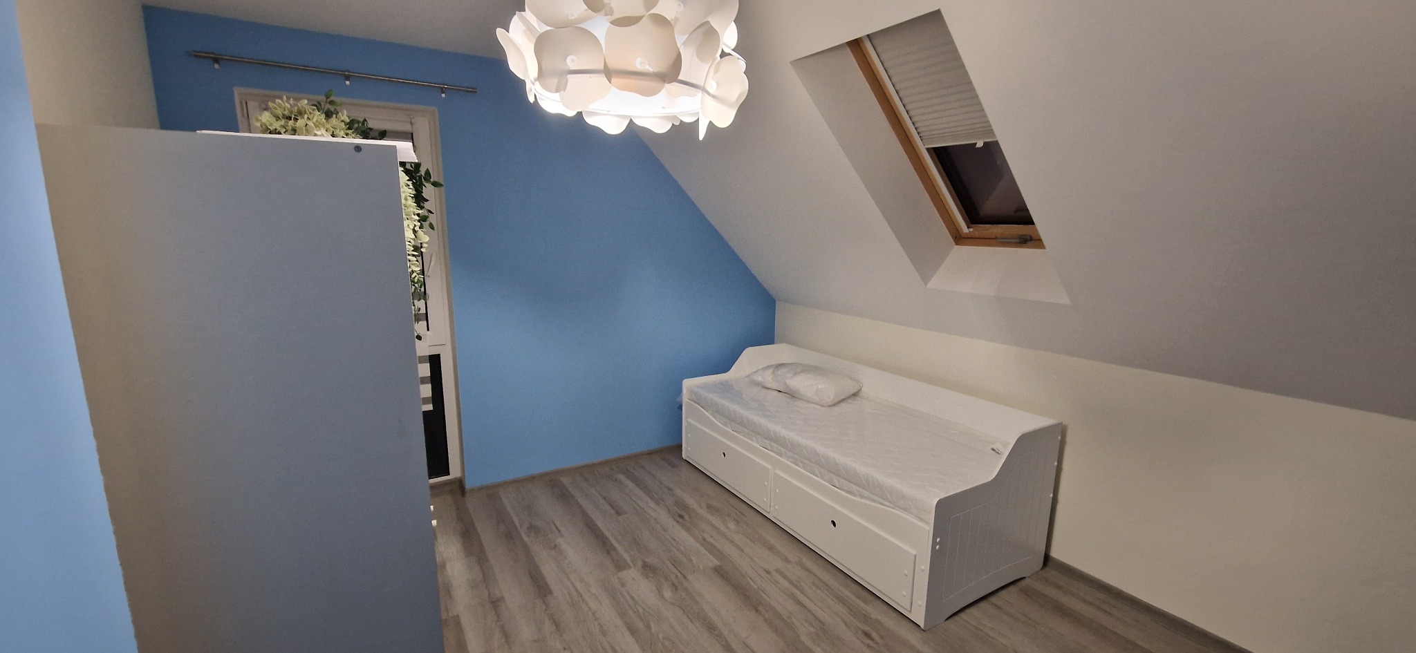 Pokój na poddaszu po odświeżeniu: błękitna ściana, białe meble (szafa, łóżko z szufladami), okno dachowe i designerska lampa. Jasne panele podłogowe.