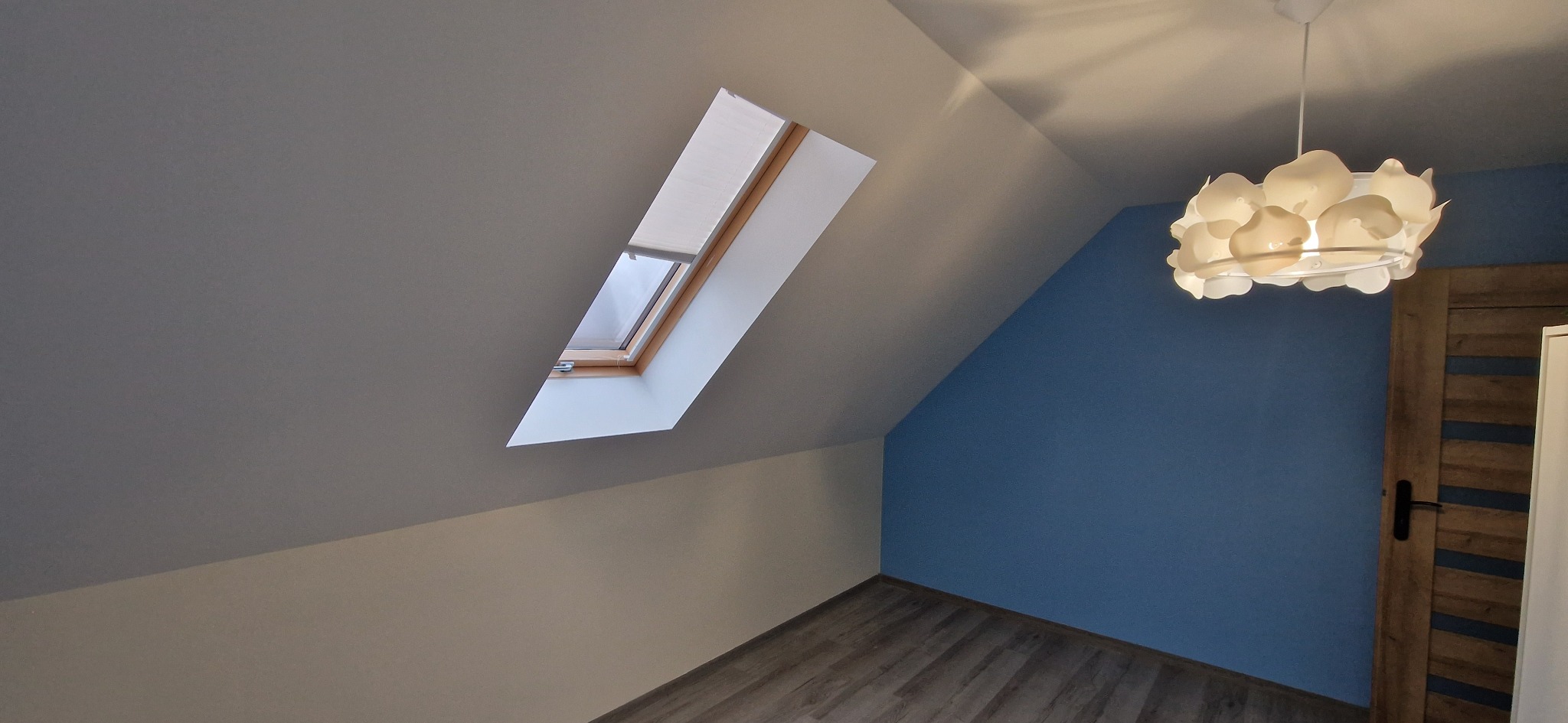 Pokój na poddaszu po remoncie: okno dachowe z roletą, ściany pomalowane na szaro i niebiesko, drewniana podłoga, nowoczesna lampa wisząca, drzwi z drewnianymi wstawkami.