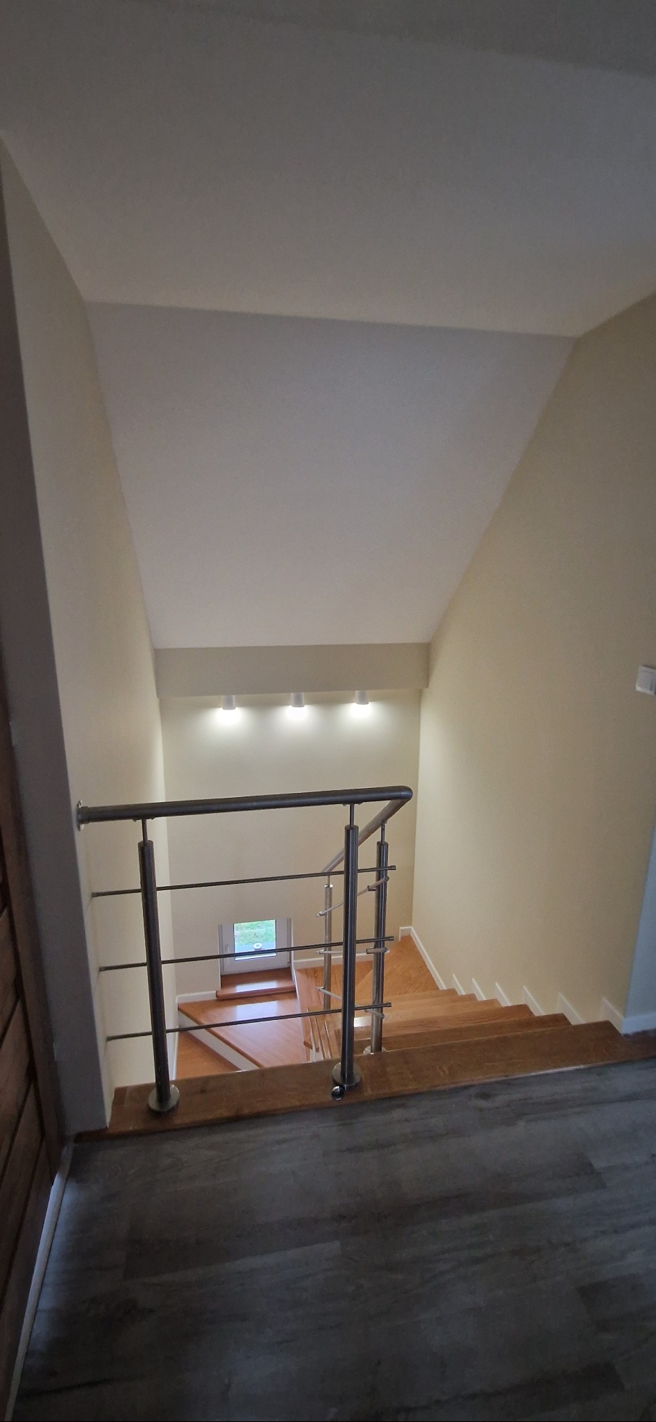 Schody drewniane z metalową balustradą, widok z góry na dół. Jasne ściany i sufit, oświetlenie punktowe. Podłoga z paneli imitujących drewno. Okno w tle.