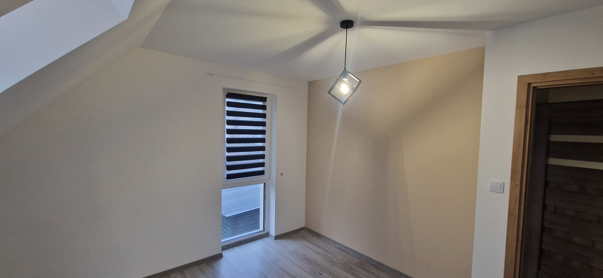 Jasne wnętrze poddasza po remoncie: kremowe ściany, okno z roletami dzień-noc, drewniana podłoga i nowoczesna lampa wisząca. Widoczny fragment drewnianych drzwi.