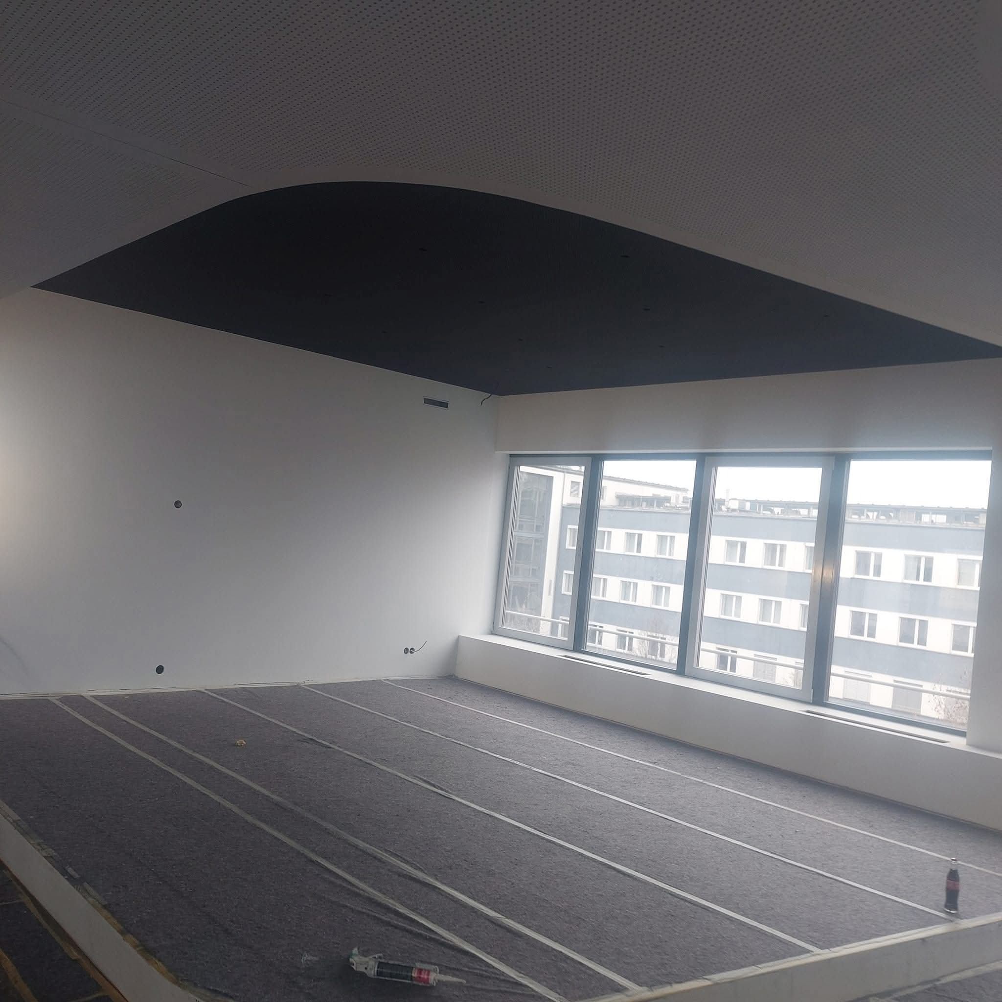 Przygotowanie podłoża pod ogrzewanie podłogowe w jasnym pokoju z dużym oknem. Widoczne listwy i mata izolacyjna. Sufit z czarnym, dekoracyjnym elementem.