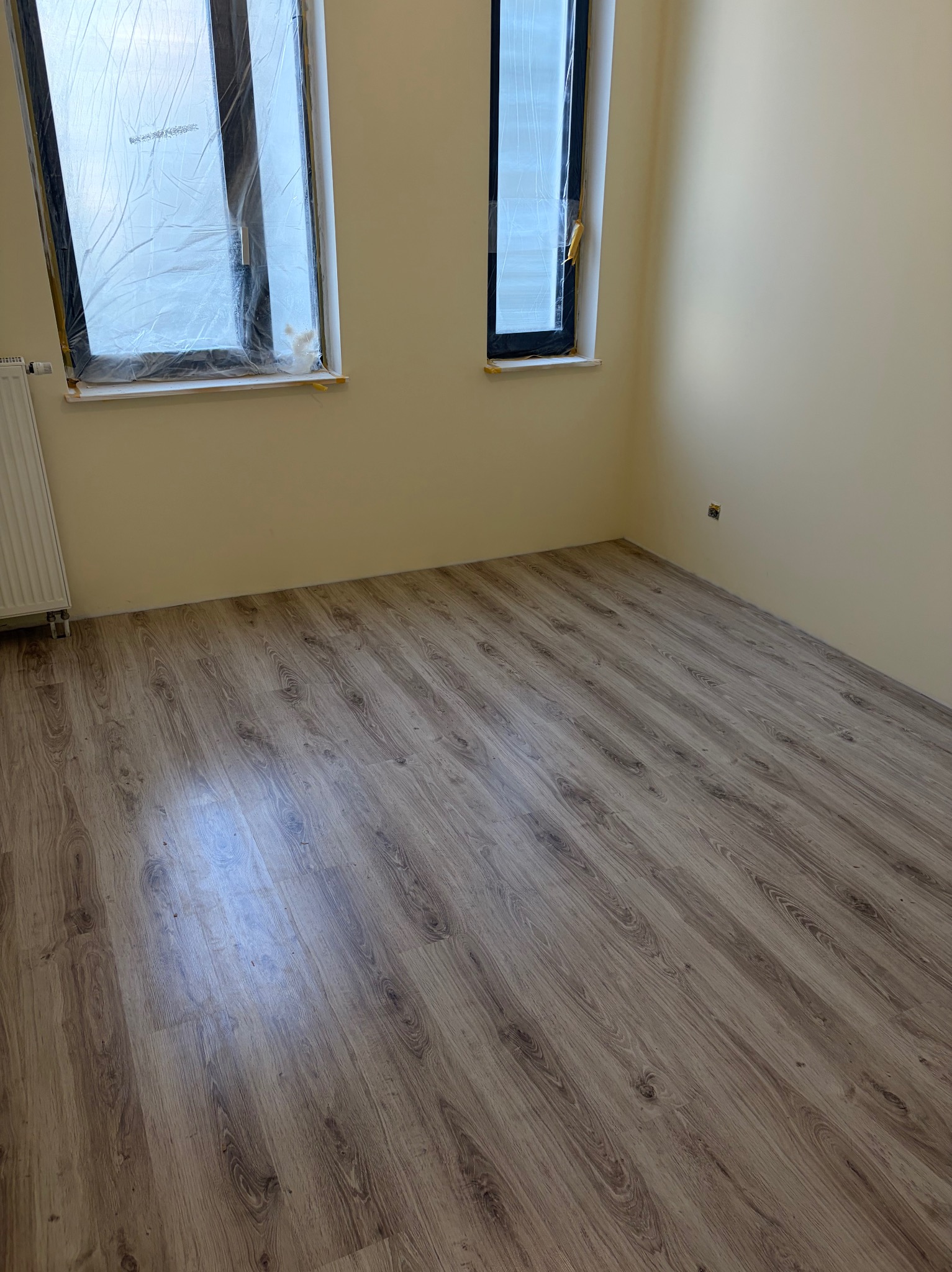 Pusty pokój po remoncie z nową podłogą z paneli i zabezpieczonymi oknami folią. Ściany pomalowane na jasny kolor. Widoczny grzejnik i gniazdko elektryczne.