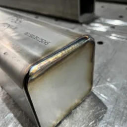 Precyzyjne spawy na metalowym profilu kwadratowym, widoczne tęczowe przebarwienia termiczne. Detal obróbki metalu na szlifowanym stole spawalniczym.