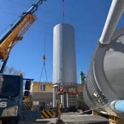 Montaż metalowego zbiornika na placu budowy przy użyciu dźwigu, z udziałem pracowników na podnośniku. Widoczne elementy konstrukcyjne i budynek w tle.