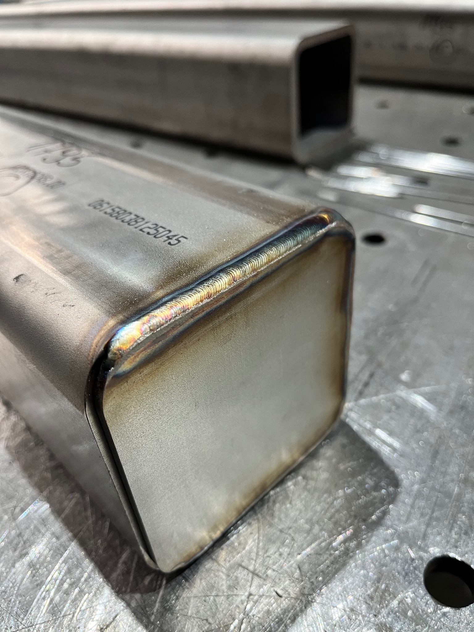 Precyzyjne spawy na metalowym profilu kwadratowym, widoczne tęczowe przebarwienia termiczne. Detal obróbki metalu na szlifowanym stole spawalniczym.