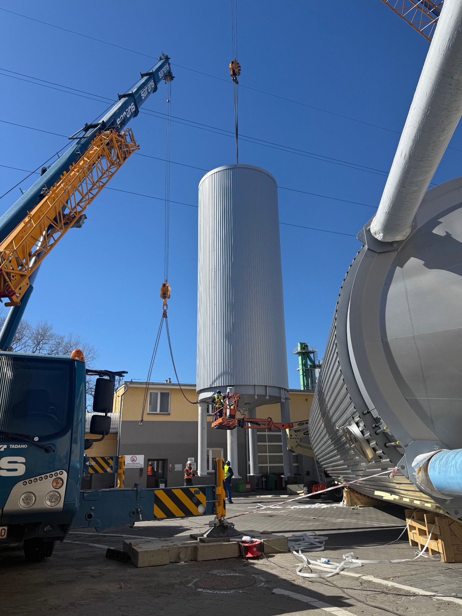 Montaż metalowego zbiornika na placu budowy przy użyciu dźwigu, z udziałem pracowników na podnośniku. Widoczne elementy konstrukcyjne i budynek w tle.