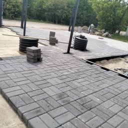 Bmw solidbruk - Szara kostka brukowa ułożona we wzór jodełki, widoczne czarne słupki konstrukcji i przygotowane materiały. Częściowo ułożona nawierzchnia, piasek i obrzeża.