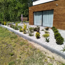 Nowoczesny ogród z obrzeżami z kostki, białym żwirem i różnorodną roślinnością na tle budynku z drewnianą elewacją i szarym brukiem.