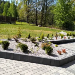 Ogród z rabatami wykończonymi szarą kostką, wypełnionymi białym grysem i różnorodną roślinnością. W tle trawnik i ogrodzenie z drewnianych paneli.