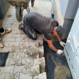 Bmw solidbruk - Mężczyzna w rękawicach układa kostkę brukową przy białej skrzynce elektrycznej, obok taczka i fragment ułożonej nawierzchni z kostki. Widoczne czarne podłoże.