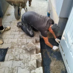 Mężczyzna w rękawicach układa kostkę brukową przy białej skrzynce elektrycznej, obok taczka i fragment ułożonej nawierzchni z kostki. Widoczne czarne podłoże.