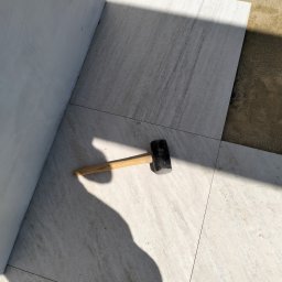 Bmw solidbruk - Układanie jasnych płyt chodnikowych przy ścianie. Gumowy młotek leży na powierzchni. Częściowo widoczne stopy w obuwiu roboczym.