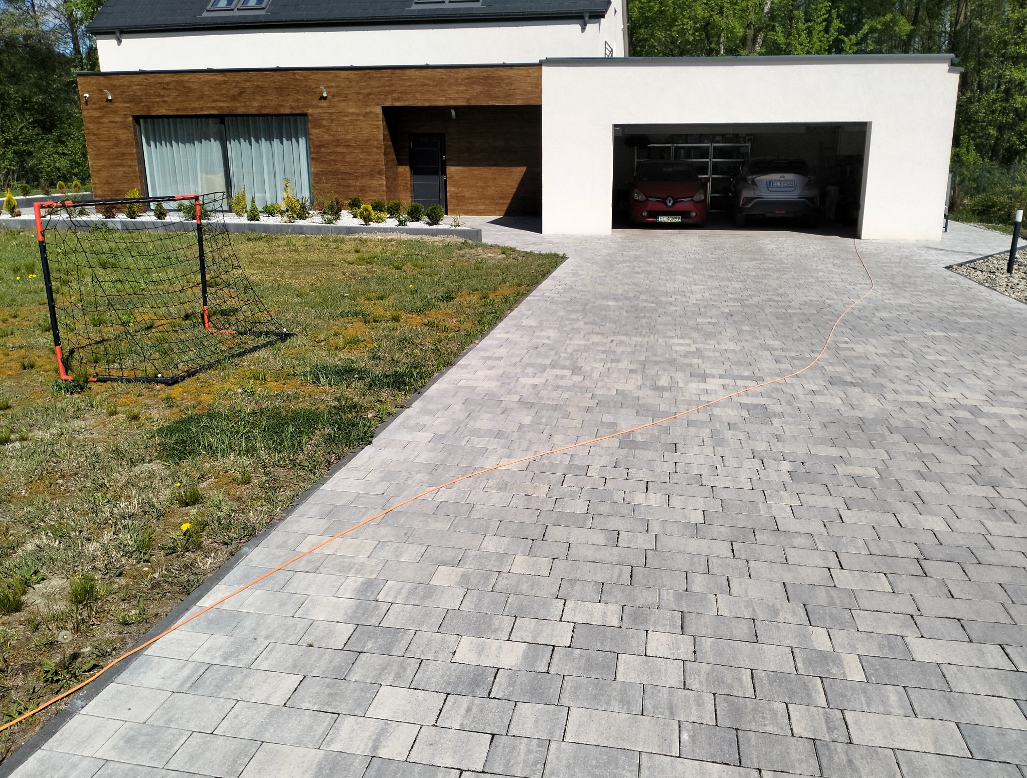 Nowoczesny podjazd z szarej kostki brukowej prowadzący do garażu dwustanowiskowego. Na trawniku bramka do piłki nożnej. Widok na dom z elementami drewnianymi.