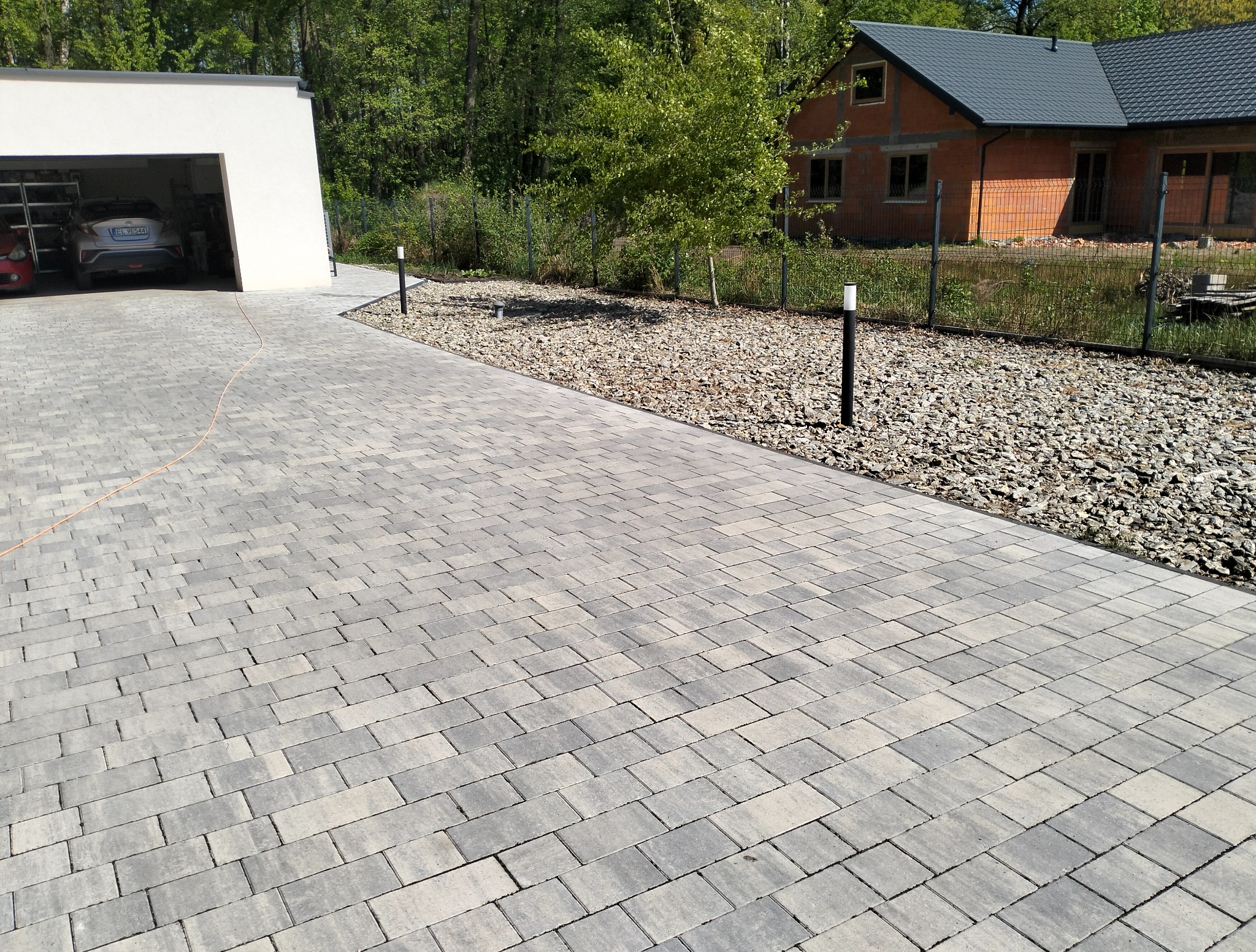 Szara kostka brukowa ułożona na podjeździe do garażu, obok rabata z kamieniami i lampami ogrodowymi. W tle dom w trakcie budowy.
